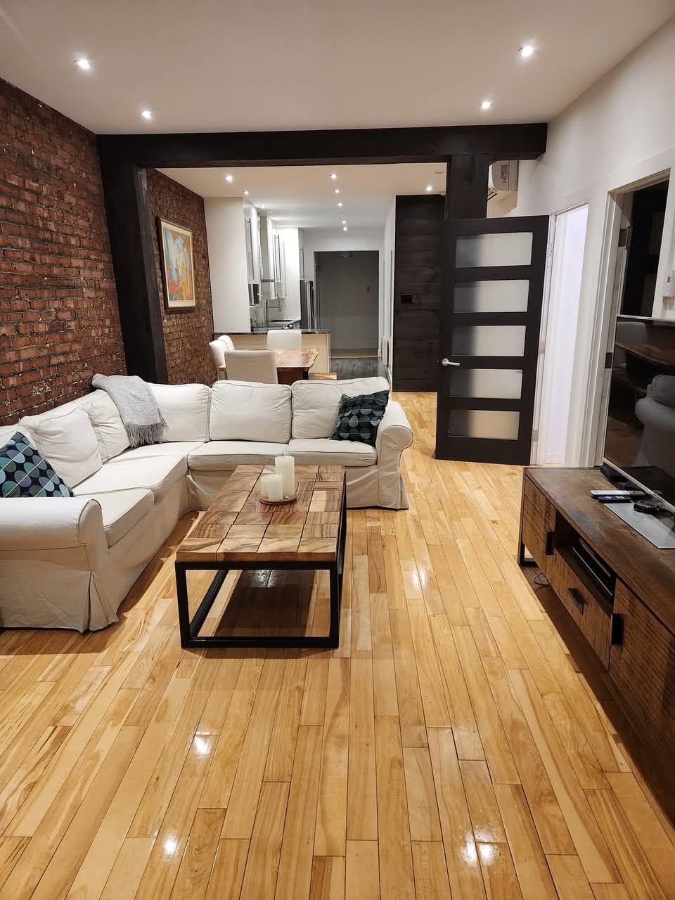 Salon d'appartement montréalais avec plancher de bois franc poli, mur de brique apparente et cuisine ouverte à l'arrière