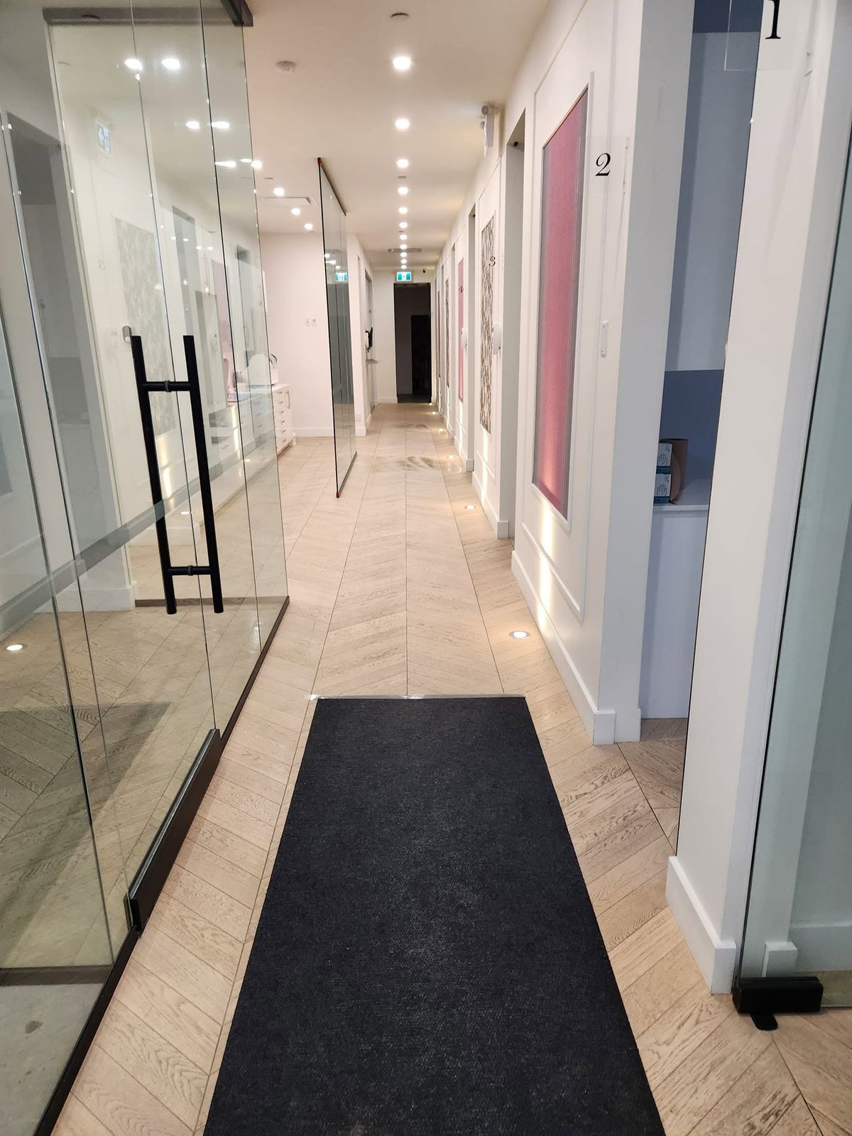 Corridor de clinique avec salles de traitement numérotées, cloisons vitrées et plancher de tuile posé en chevron